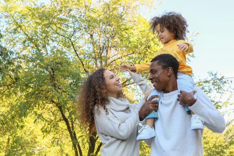 Family of three in a park: Child on father's shoulders, mother smiling, sunny day.