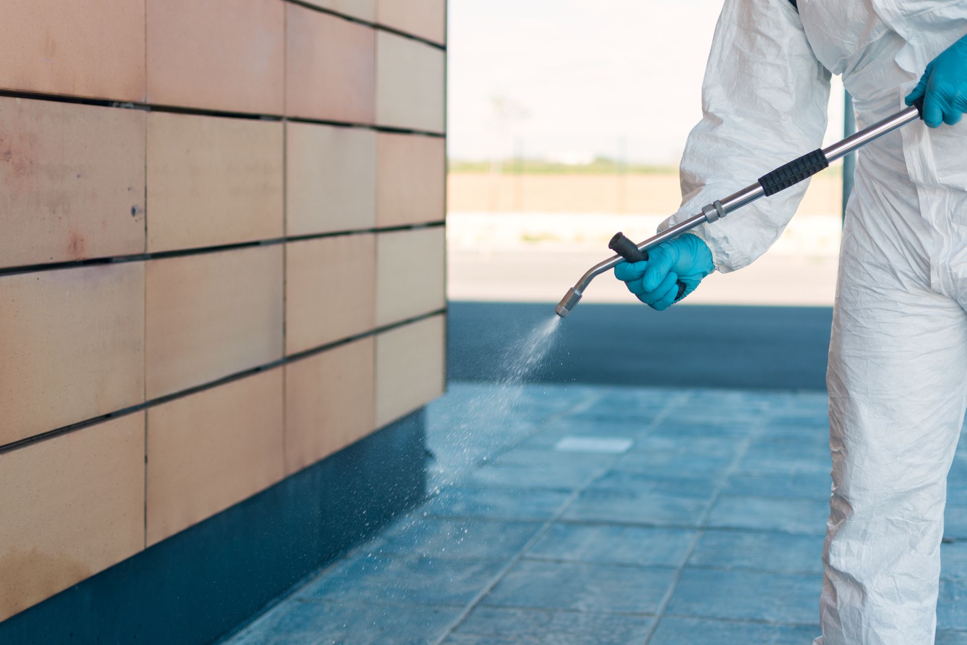 Person in protective suit spraying a surface with a silver nozzle, next to a brick wall.
