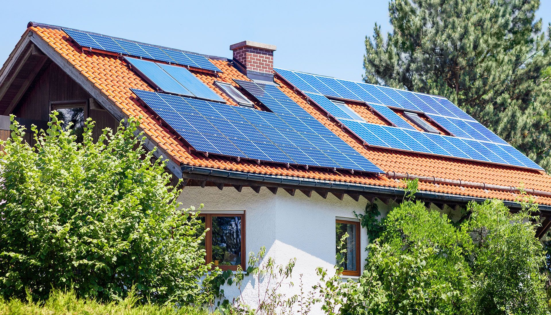 Solar panels on a red-tiled roof of a house with white exterior walls and surrounding green trees.