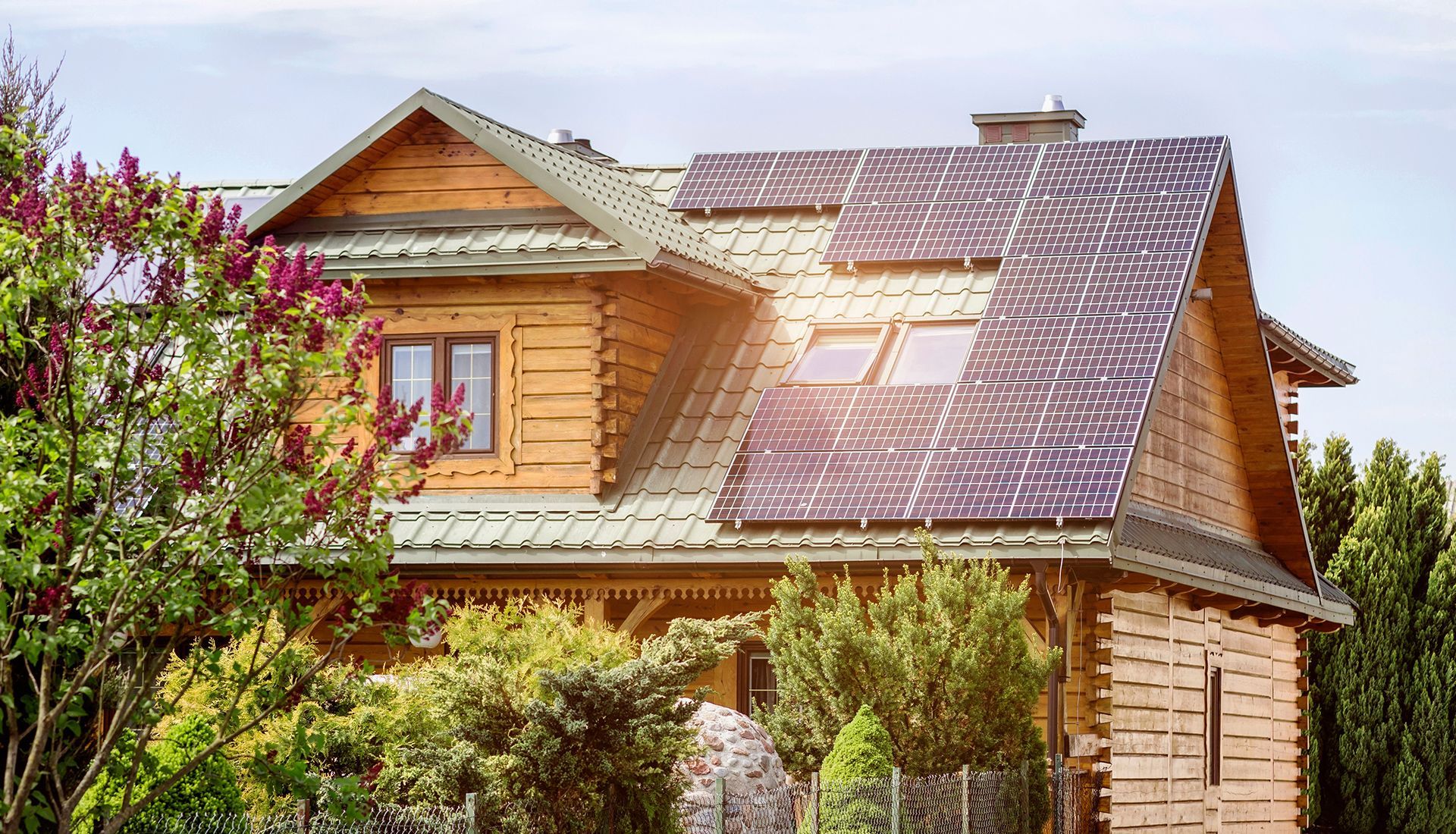Wooden house with solar panels on the roof, surrounded by trees and greenery under a bright sky.