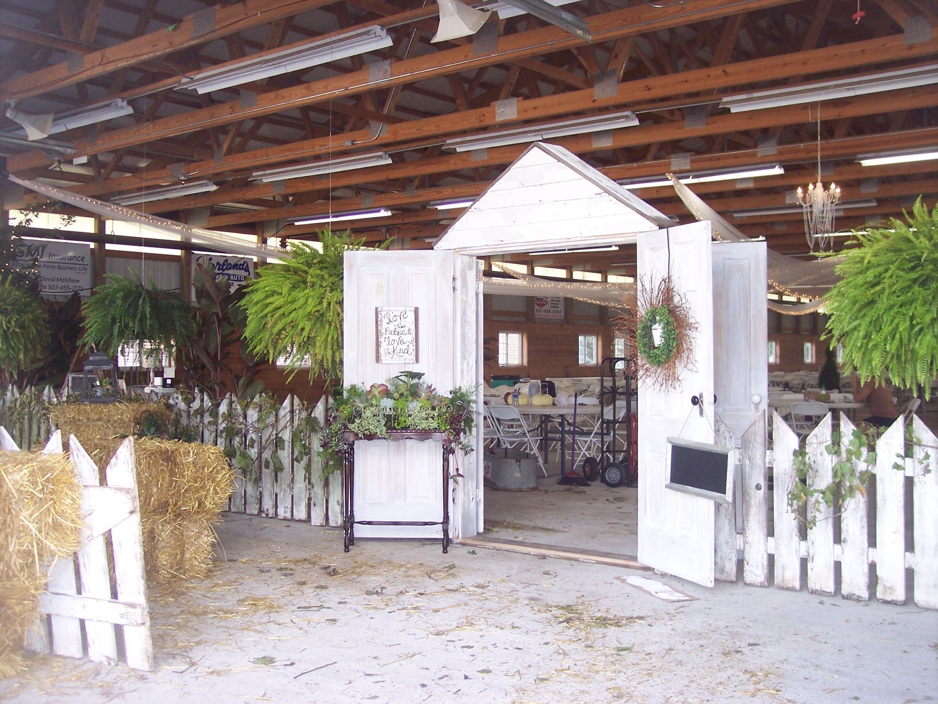 A rustic indoor event space featuring a white wooden faux-cottage entrance, hay bales, and greenery under wood rafters.