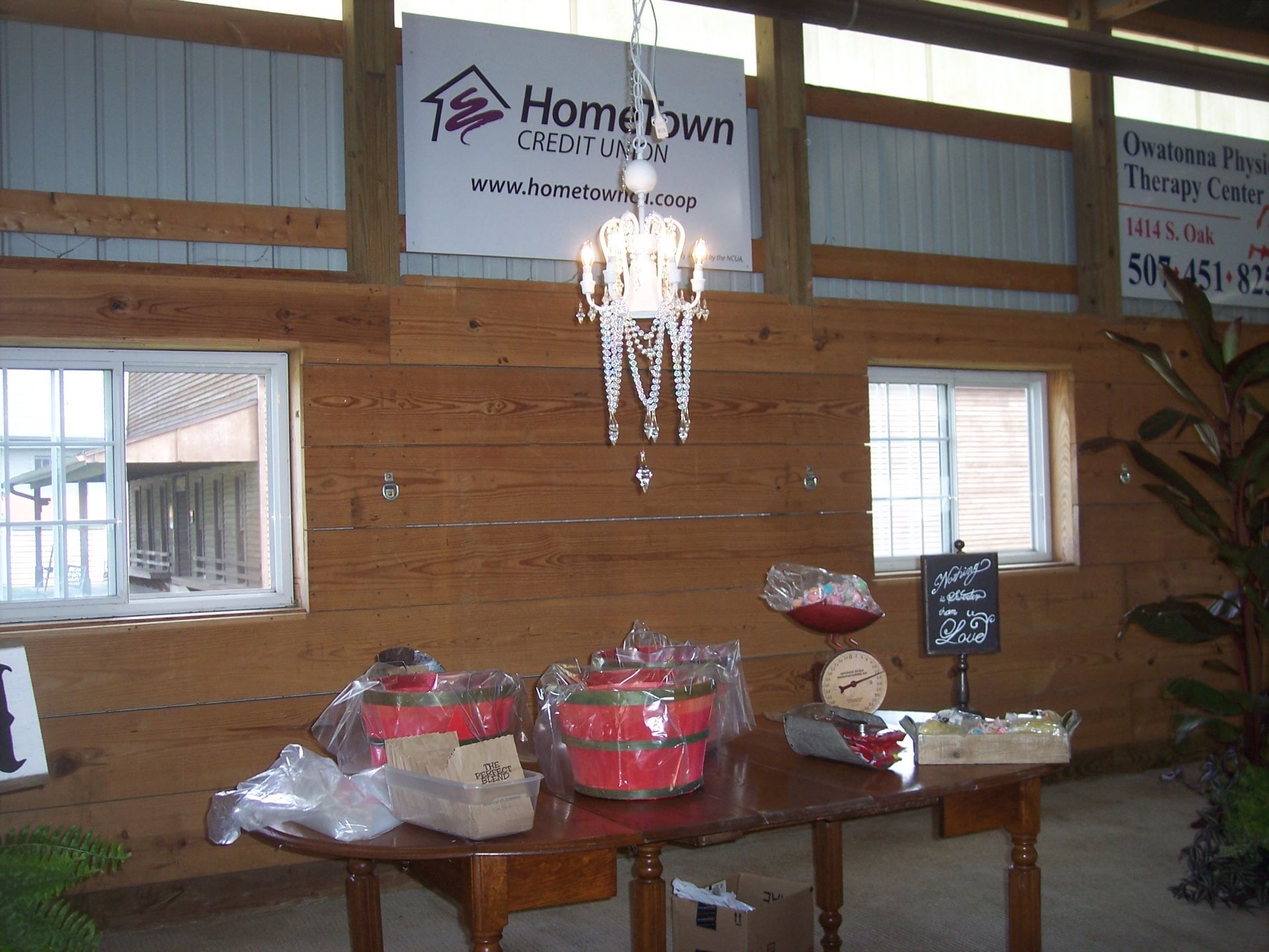 A rustic wooden interior with a table holding red gift baskets and a white chandelier hanging above, below signage.