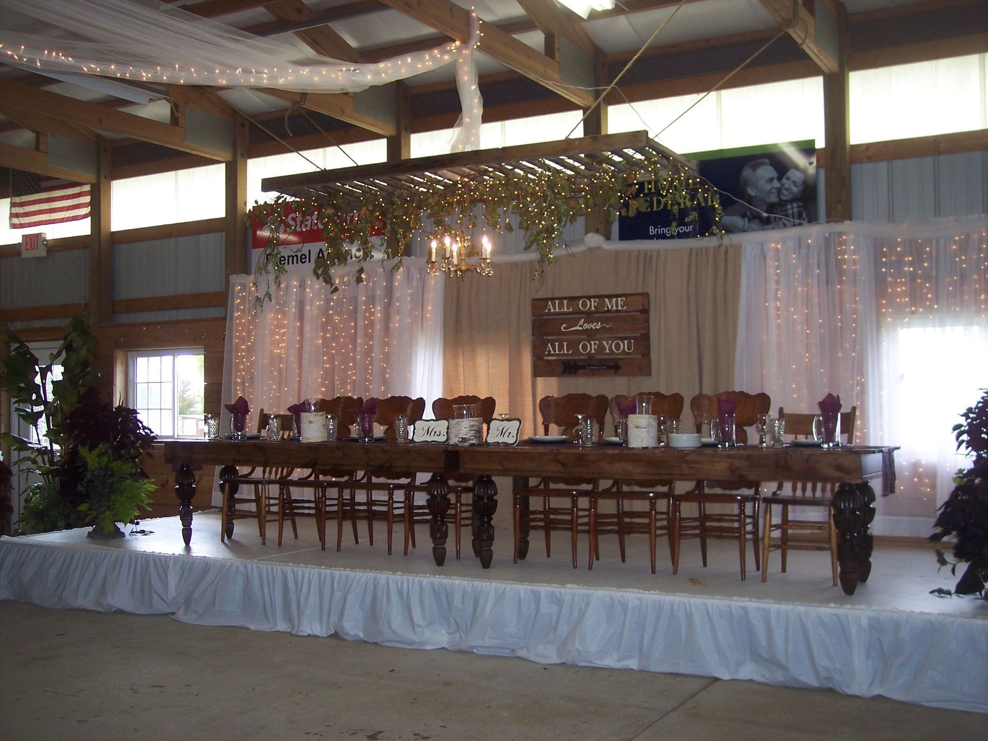 A long wedding head table on a raised platform, decorated with string lights, fabric drapes, and a rustic wooden sign.