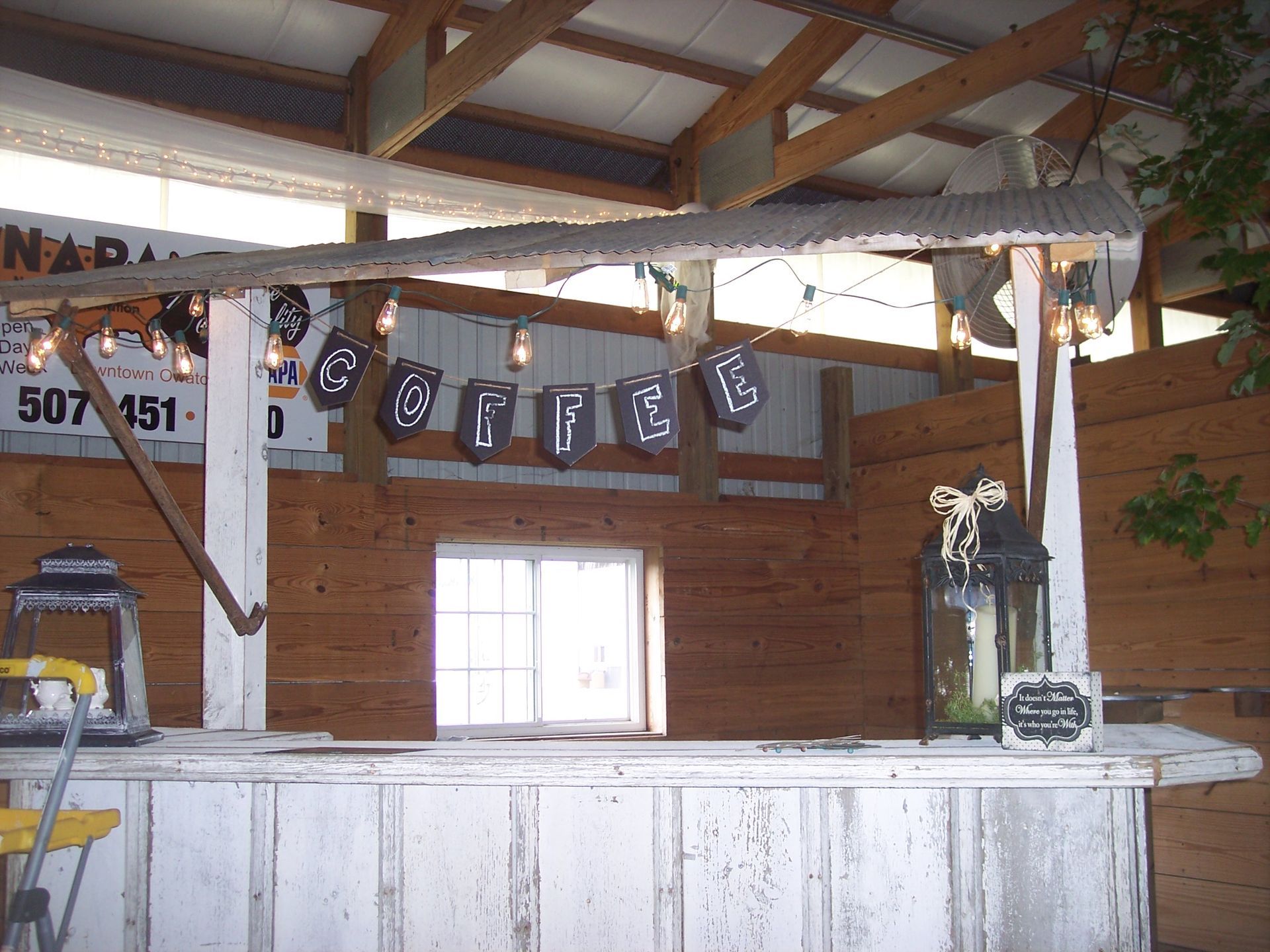 A rustic, white wooden coffee bar with a corrugated metal roof, string lights, and a hanging 