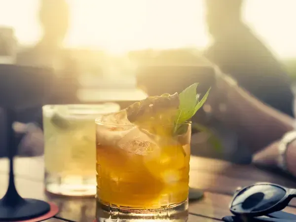 Cocktail in a glass with ice and garnish, on a wooden table outdoors. Sunshine in the background.