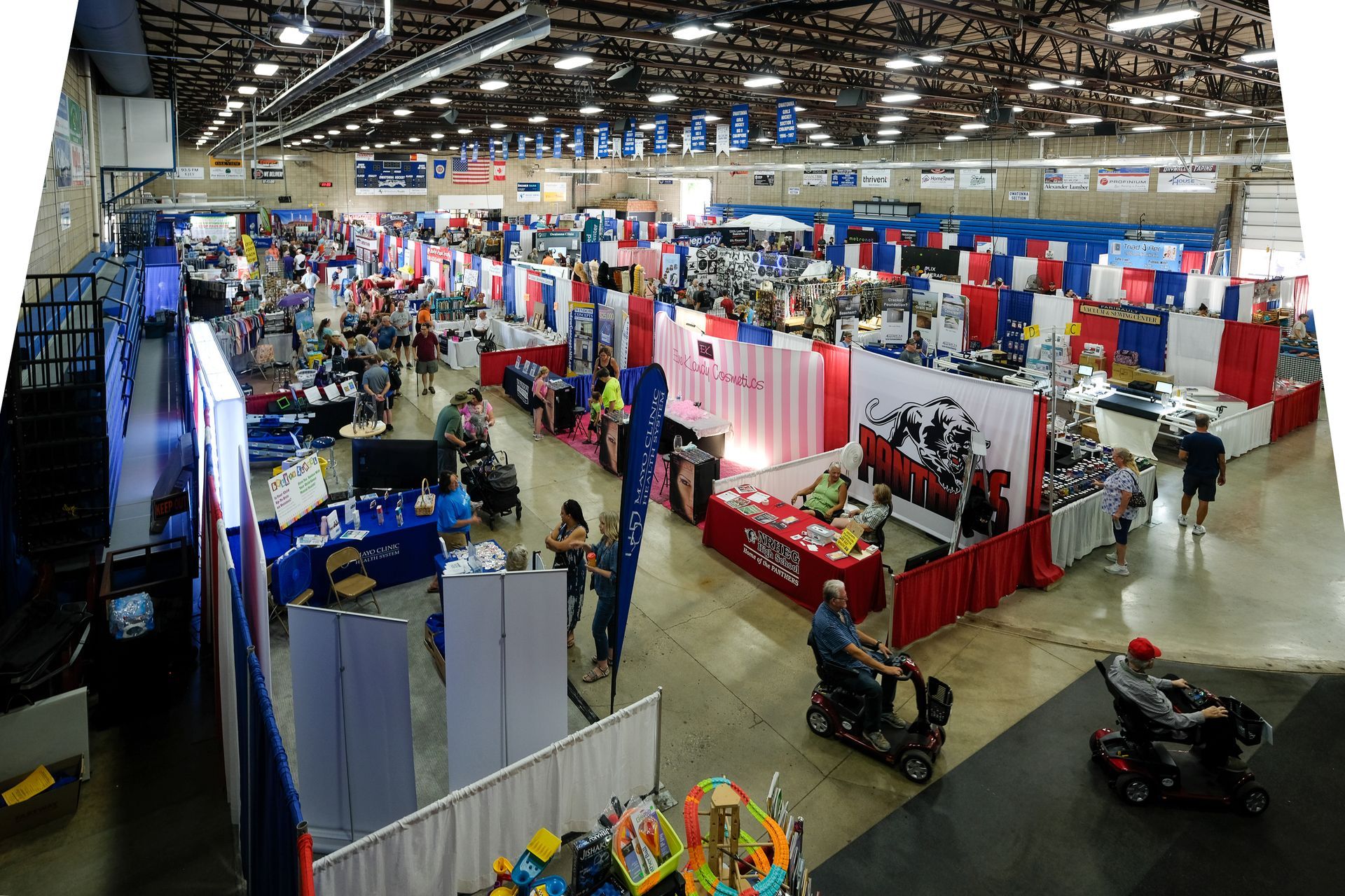 Large event hall with booths, people browsing. Red, white, and blue decor. Two people use mobility devices.