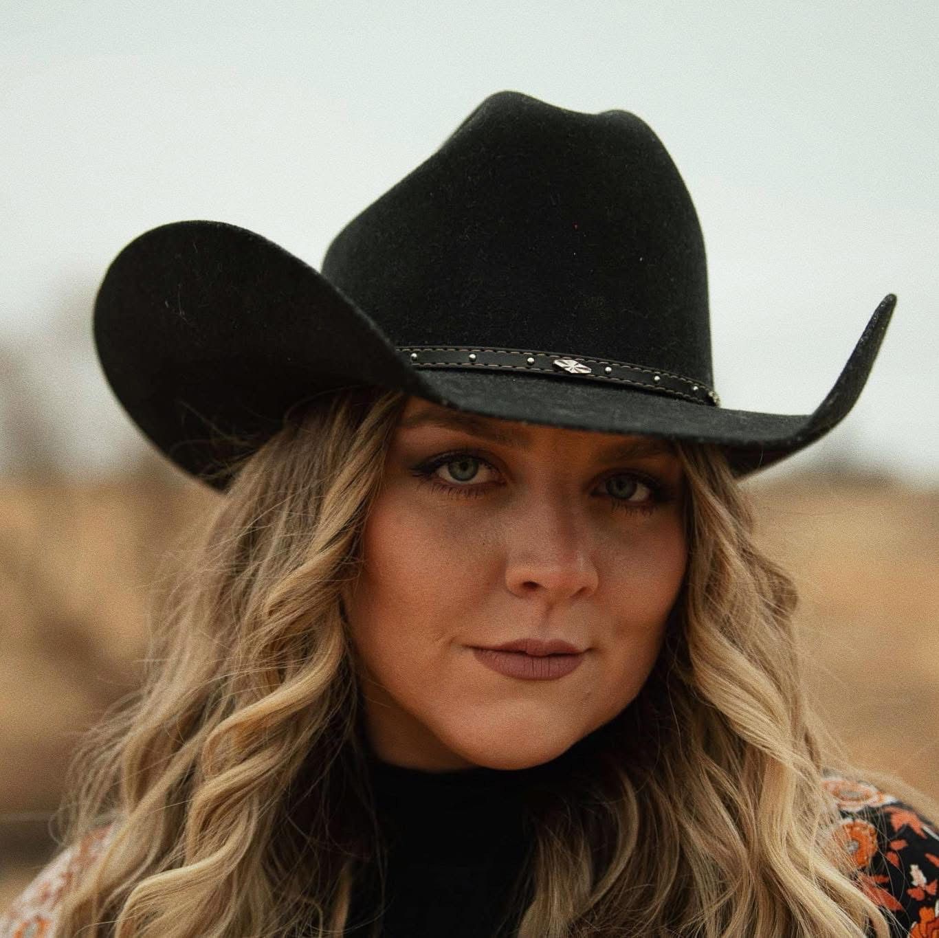 Cori Kennedy in black cowboy hat, long wavy blonde hair, looking towards the camera. Outdoor setting.