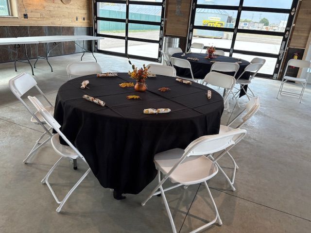 A round table with a black cloth and white folding chairs in a rustic, industrial-style room with large glass doors.