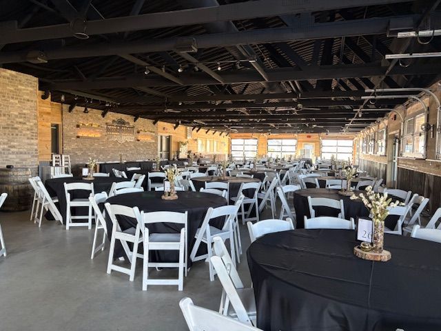An indoor event space set up for a banquet with black-clothed round tables and white folding chairs under a black ceiling.