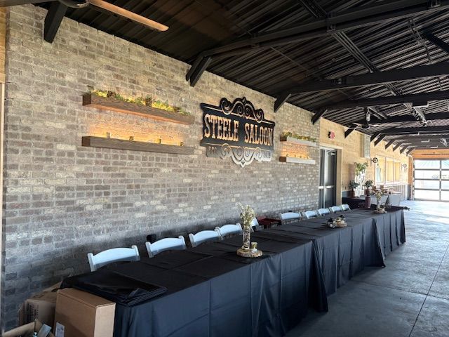 Brick-walled Steele Saloon interior with long tables, white chairs, and a garage door.