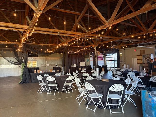 Event space with round black tablecloths, white folding chairs, string lights, and draped black fabric.
