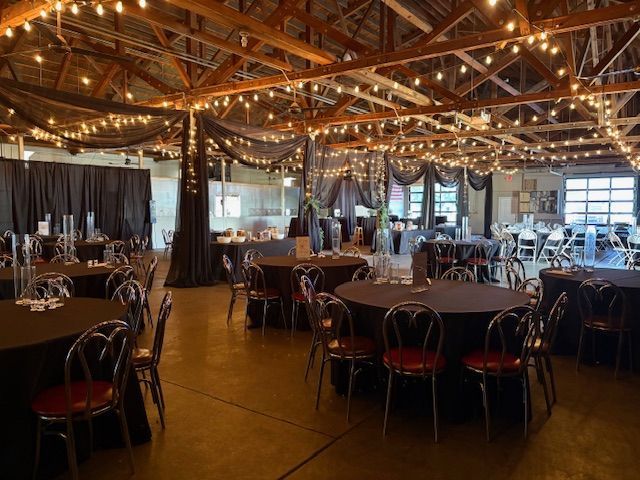 Event space with round tables, dark linens, and string lights. Black draping hangs from the ceiling.