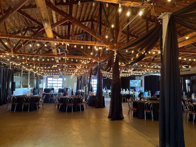 An indoor event space featuring exposed wooden beams, strings of warm lights, and tables with black linens and chairs.