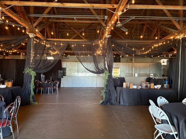 A large, rustic event space decorated with black fabric drapes, string lights, and tables set up for a gathering.