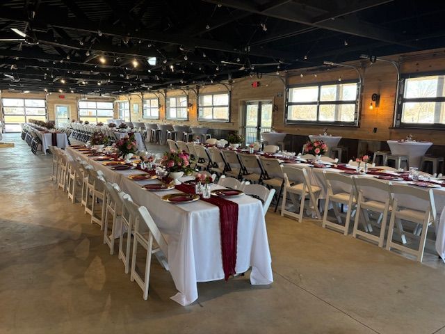 Long banquet tables with white cloths and burgundy runners, set for a formal event in a spacious venue with string lights.