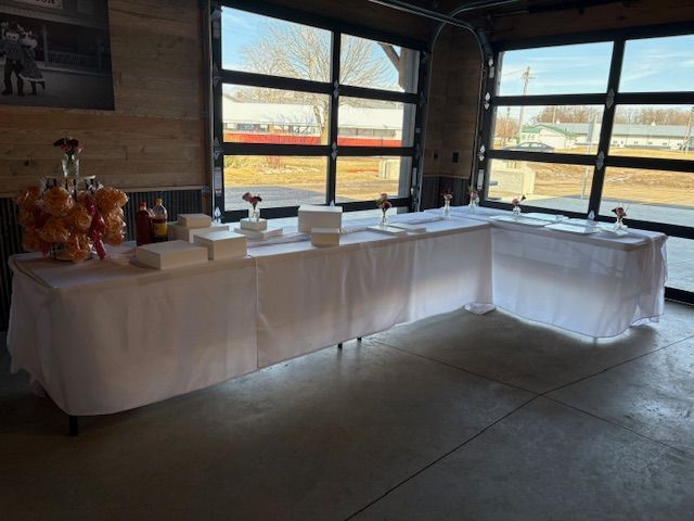 An L-shaped table with a white tablecloth, holding snacks and gift boxes in a room with large glass garage-style doors.