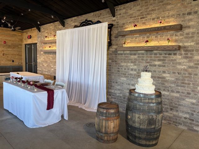 A tiered wedding cake sits atop a wooden barrel in a rustic venue with brick walls, a white backdrop, and a banquet table.