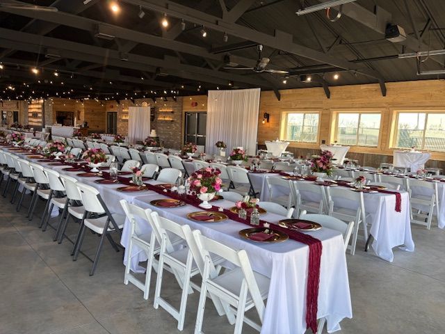 A long, banquet-style table set for a wedding in a rustic, open-plan hall with dark wooden ceilings and white chairs.