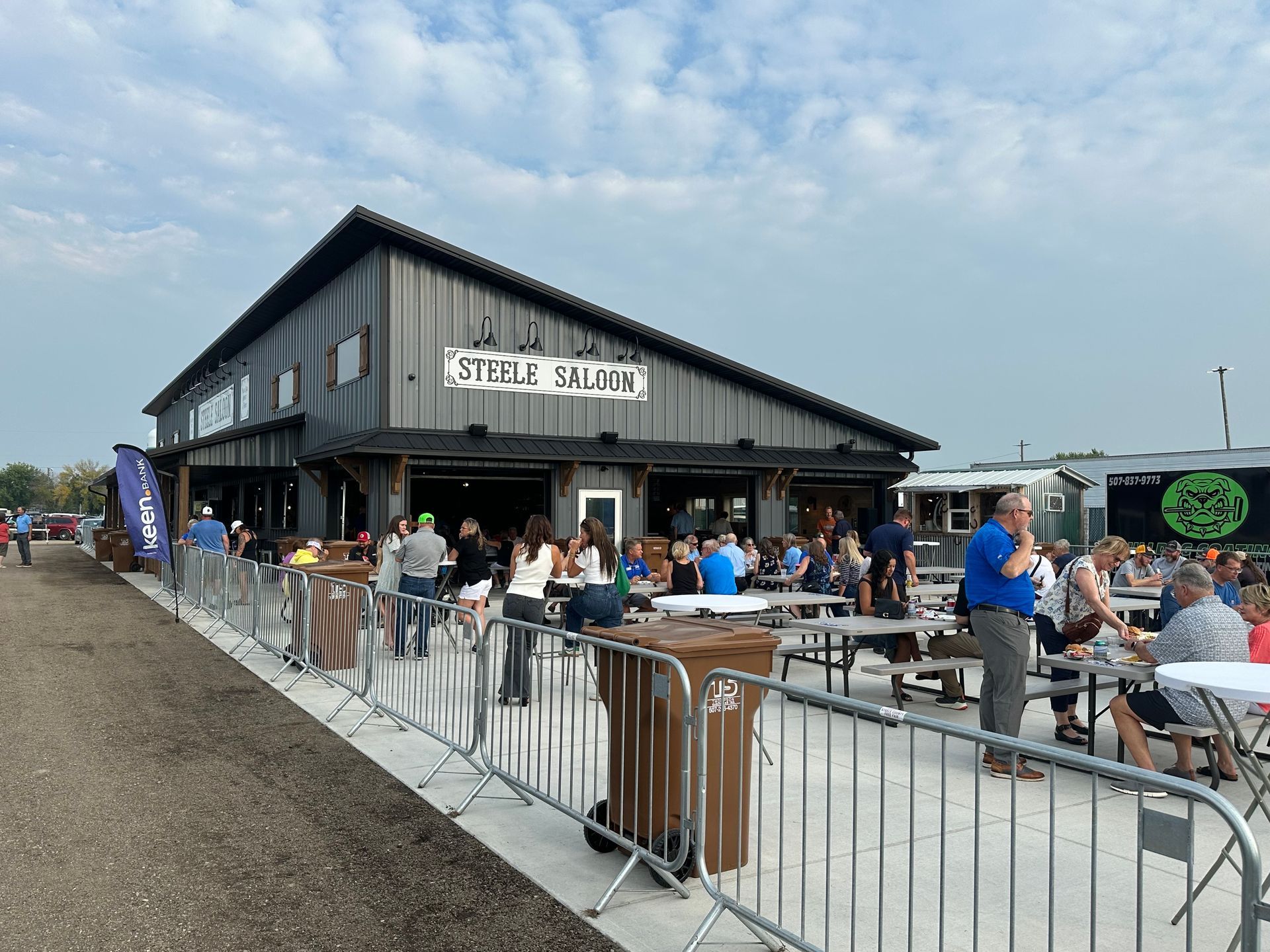 A crowd gathers on a patio outside a gray, modern metal building labeled 