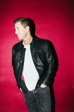 Jeremy Camp in a black leather jacket and white t-shirt poses against a solid red background, smiling and looking to the side