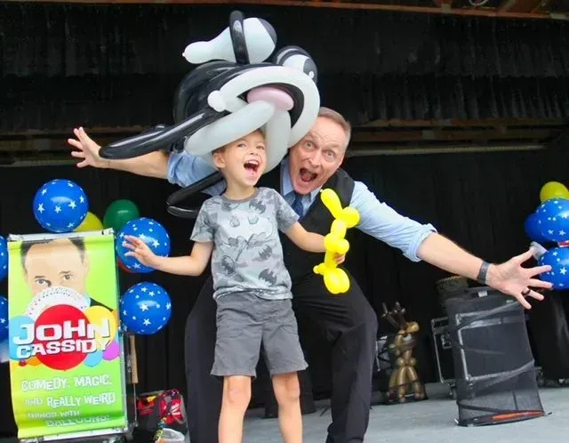 Comedy Magician John Cassidy entertaining a child with balloons on a stage, both smiling with arms outstretched.