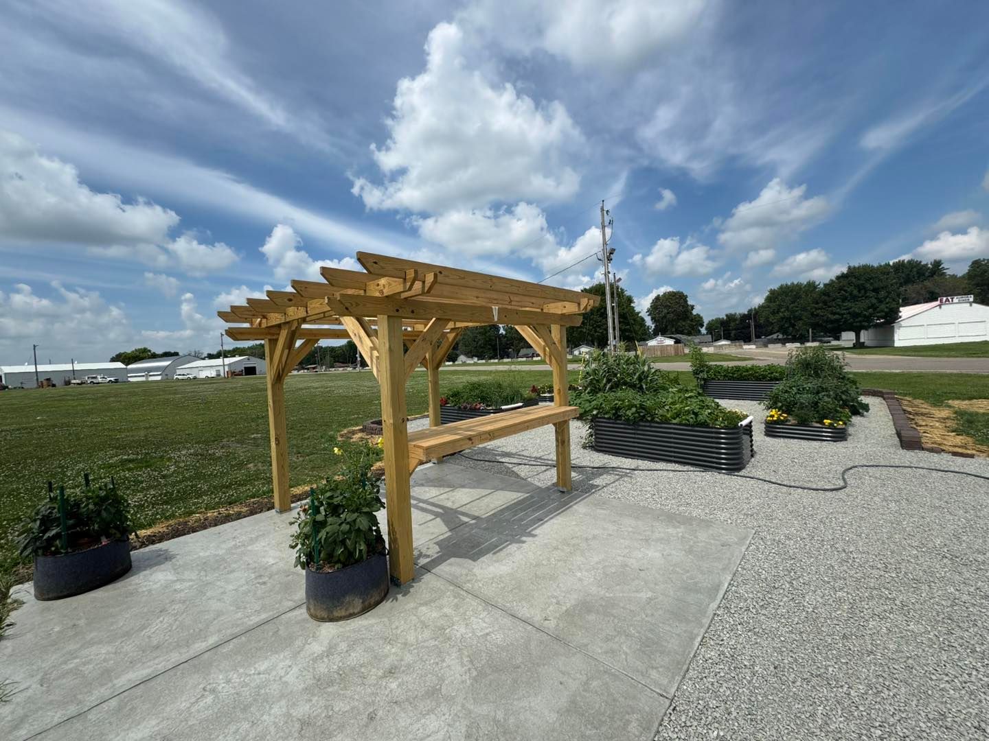 Wooden pergola with a built-in bench, next to raised garden beds on a concrete pad, under a blue sky.