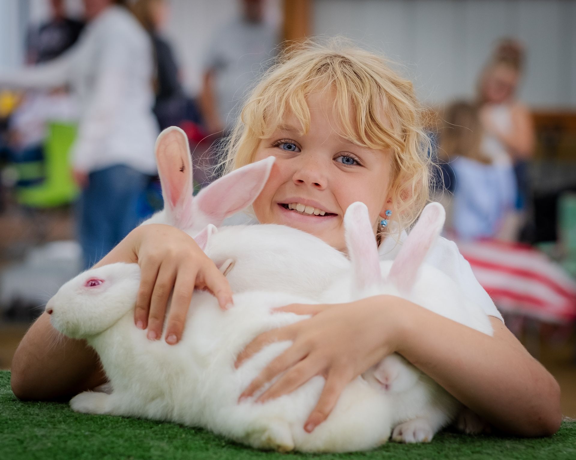 Blonde child smiles, cuddling three white rabbits on a green table at an event.