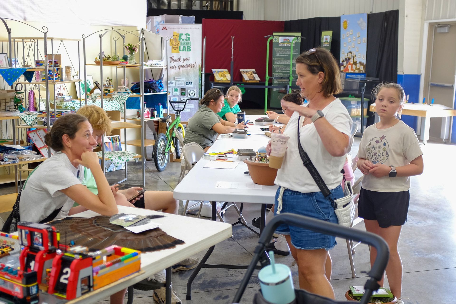 People at a table inside a building; a woman speaks while holding a drink, another sits, and a child stands nearby.
