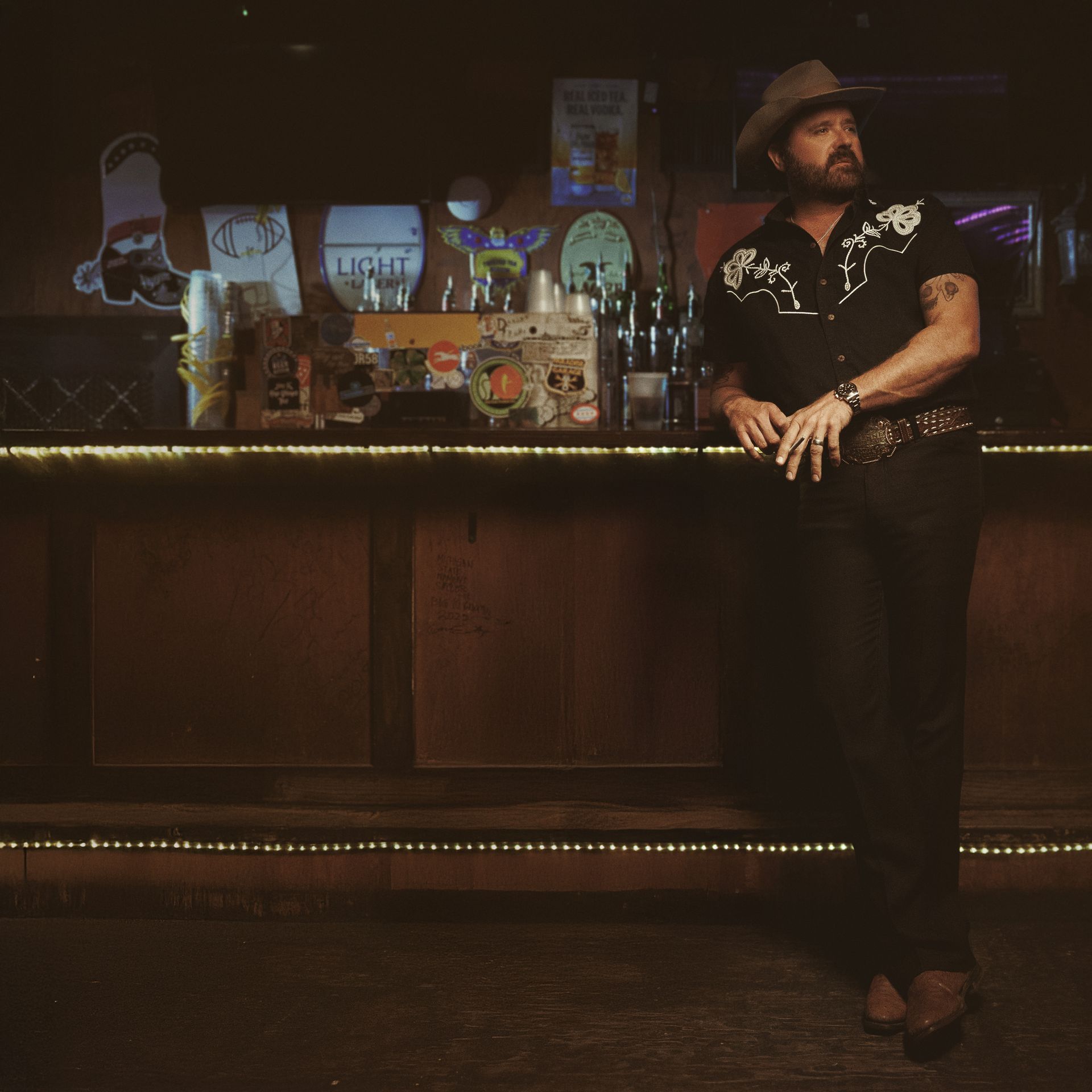 Randy Houser in cowboy hat leaning on a bar. Dark shirt, brown boots. Low-lit, bottles behind him.