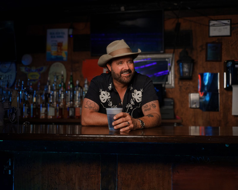 Randy Houser in a cowboy hat and embroidered shirt holding a plastic cup while leaning on a wooden bar in a tavern.