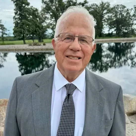 Man in gray suit and tie smiles outdoors near a body of water, trees in the background.