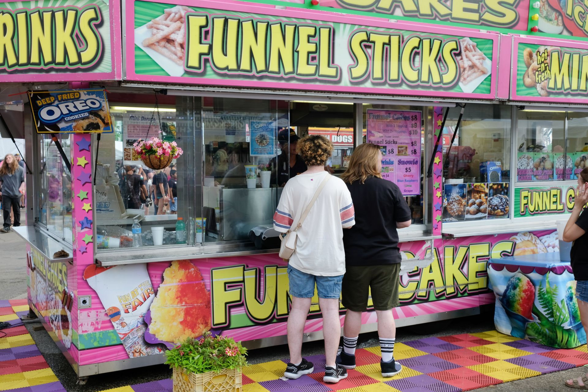 Food stand selling funnel sticks and cakes with two customers waiting.