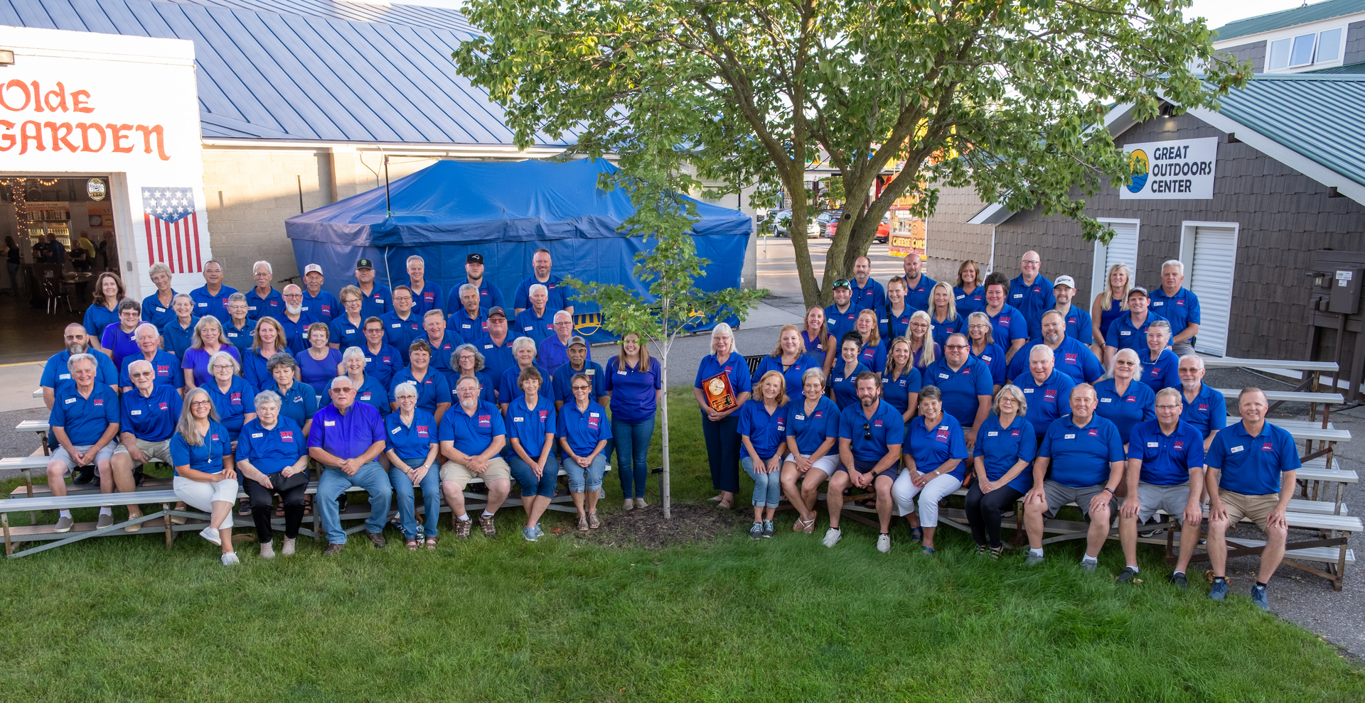 Large group of people in blue shirts posing outdoors near buildings, some seated.