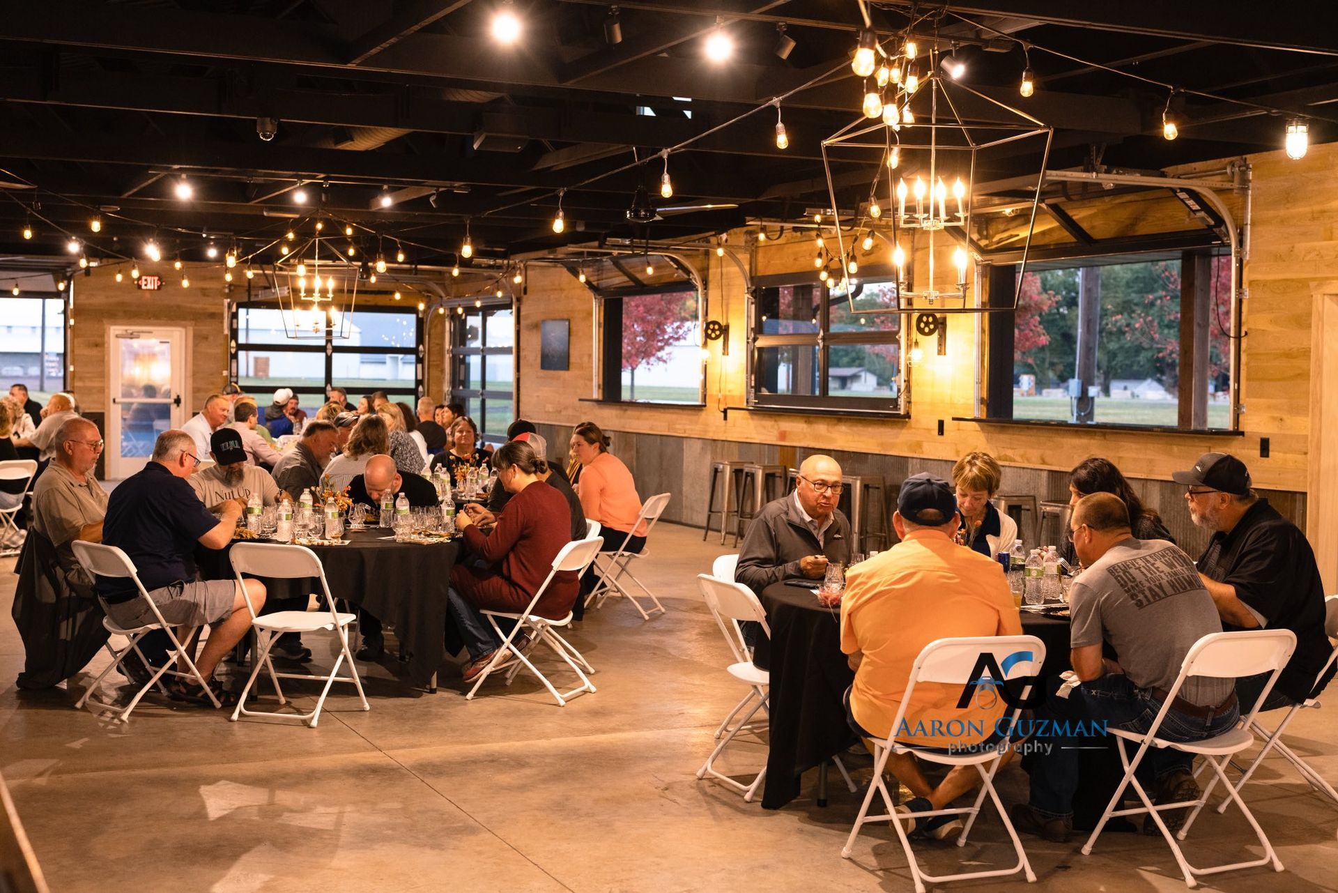 People seated at tables in a brightly lit event space with string lights and windows.