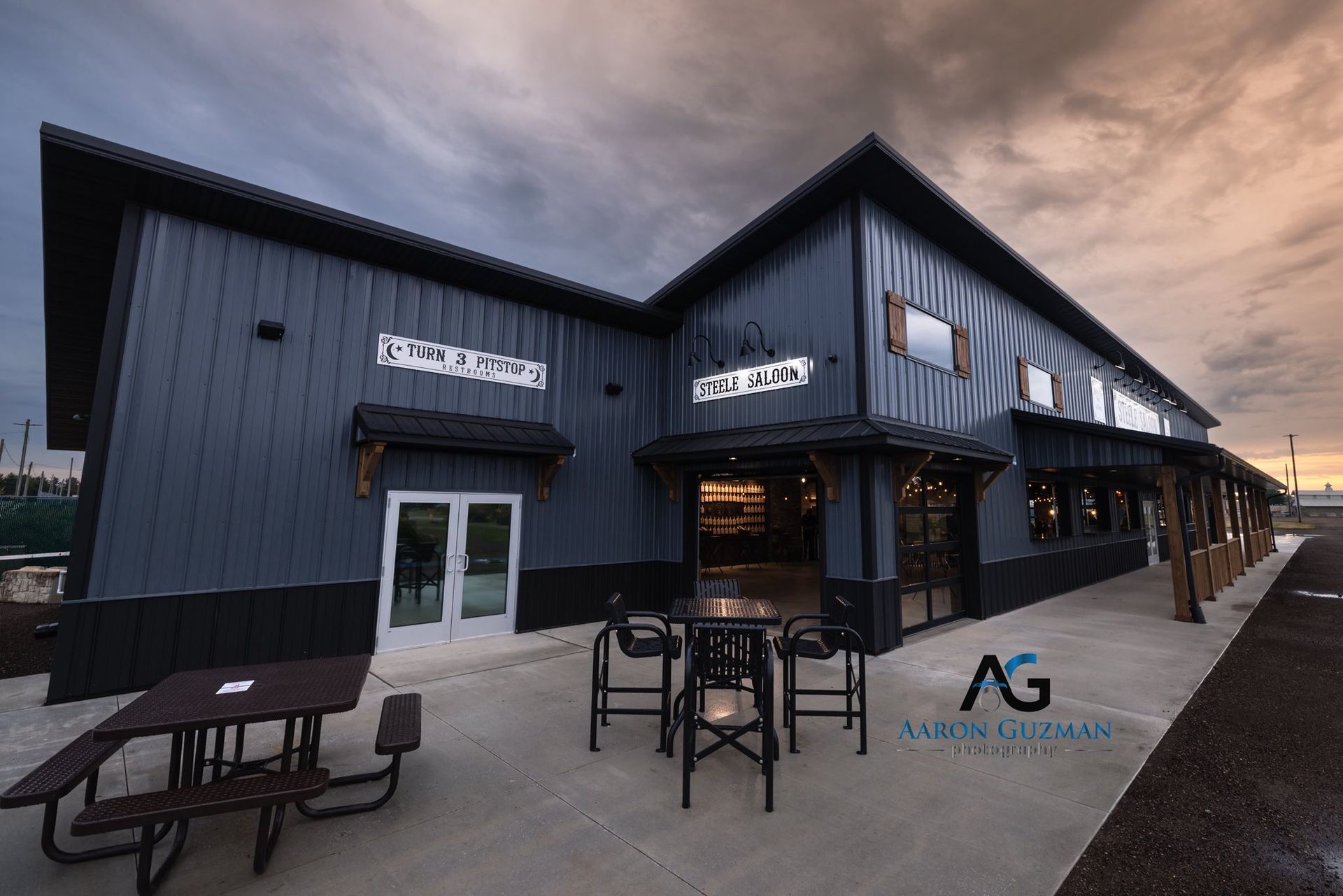 A modern dark-blue metal building with outdoor patio seating and a logo at twilight.