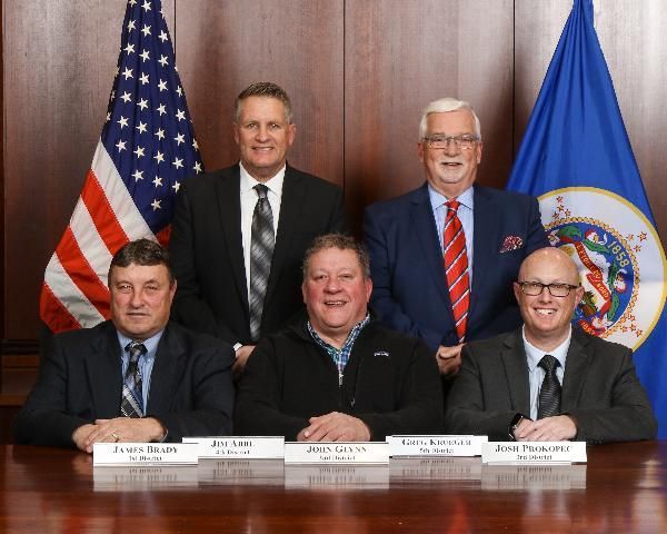 Group of men in suits posing for a photo, seated at a table with flags in the background.