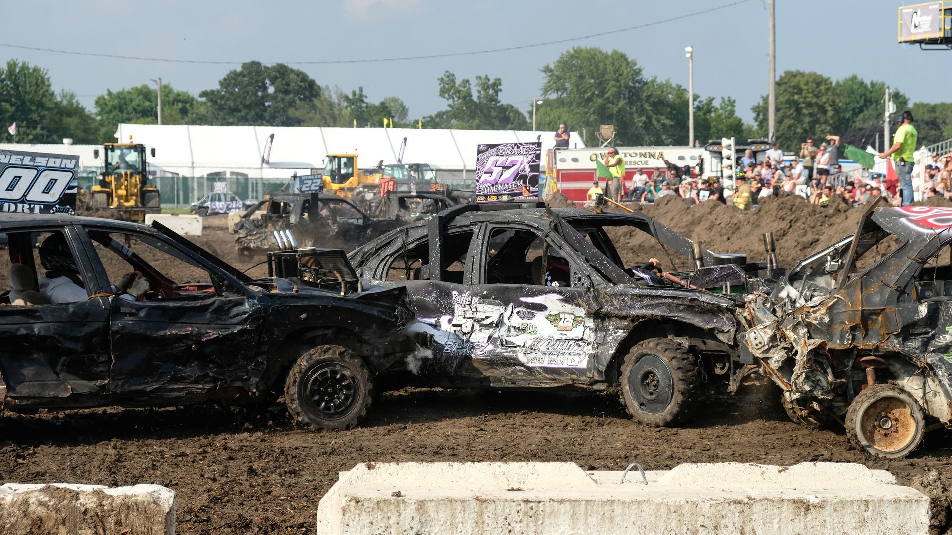 Demolition derby with three damaged cars colliding in a dirt arena, with spectators in the background.