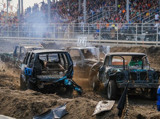 Damaged cars compete in a muddy demolition derby as a crowd watches from the stands.