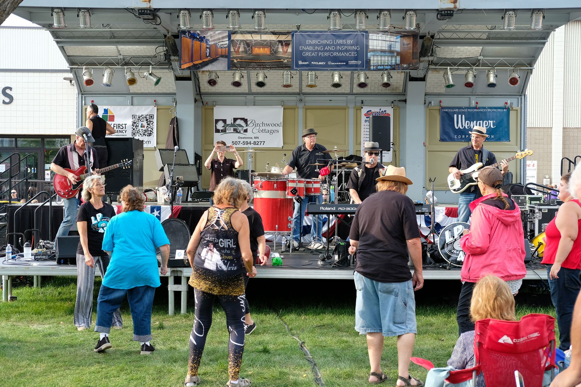 Band performing on a stage outdoors; crowd dancing on grass.