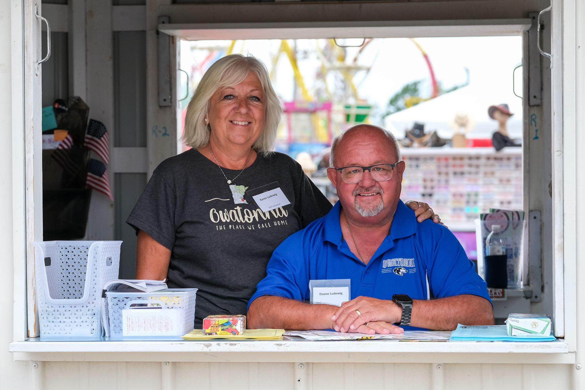 Couple smiling behind a Information Station Booth window at the fair, with a Ferris wheel in the background.