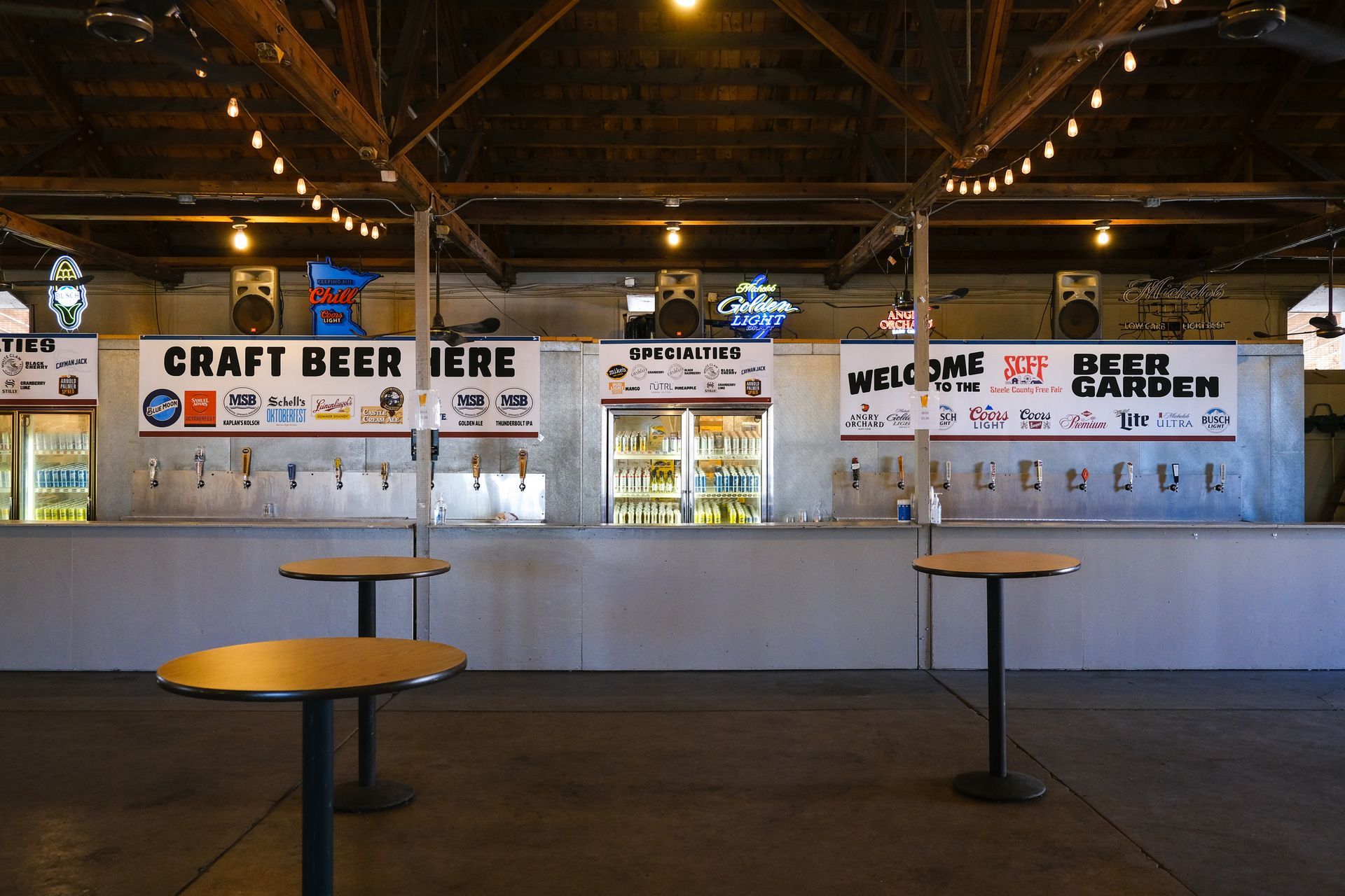 Indoor beer garden with bar, taps, signs for craft beer. Empty tables, string lights, and concrete floor.