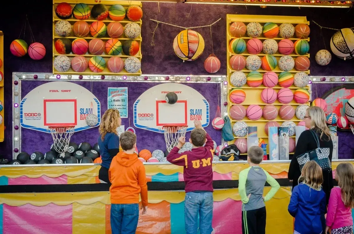 Kids playing basketball carnival game, colorful balls and prizes in the background.