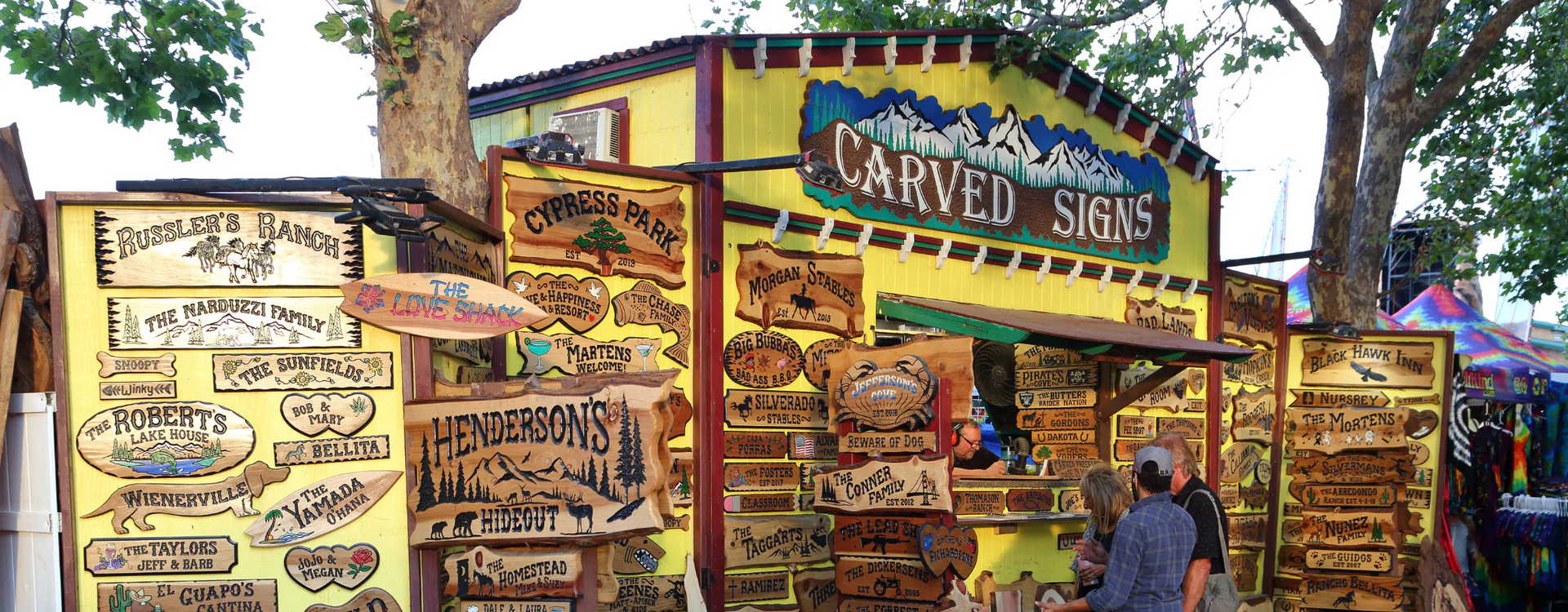 A shop selling carved signs is displayed with various signs hanging on the walls and two customers.