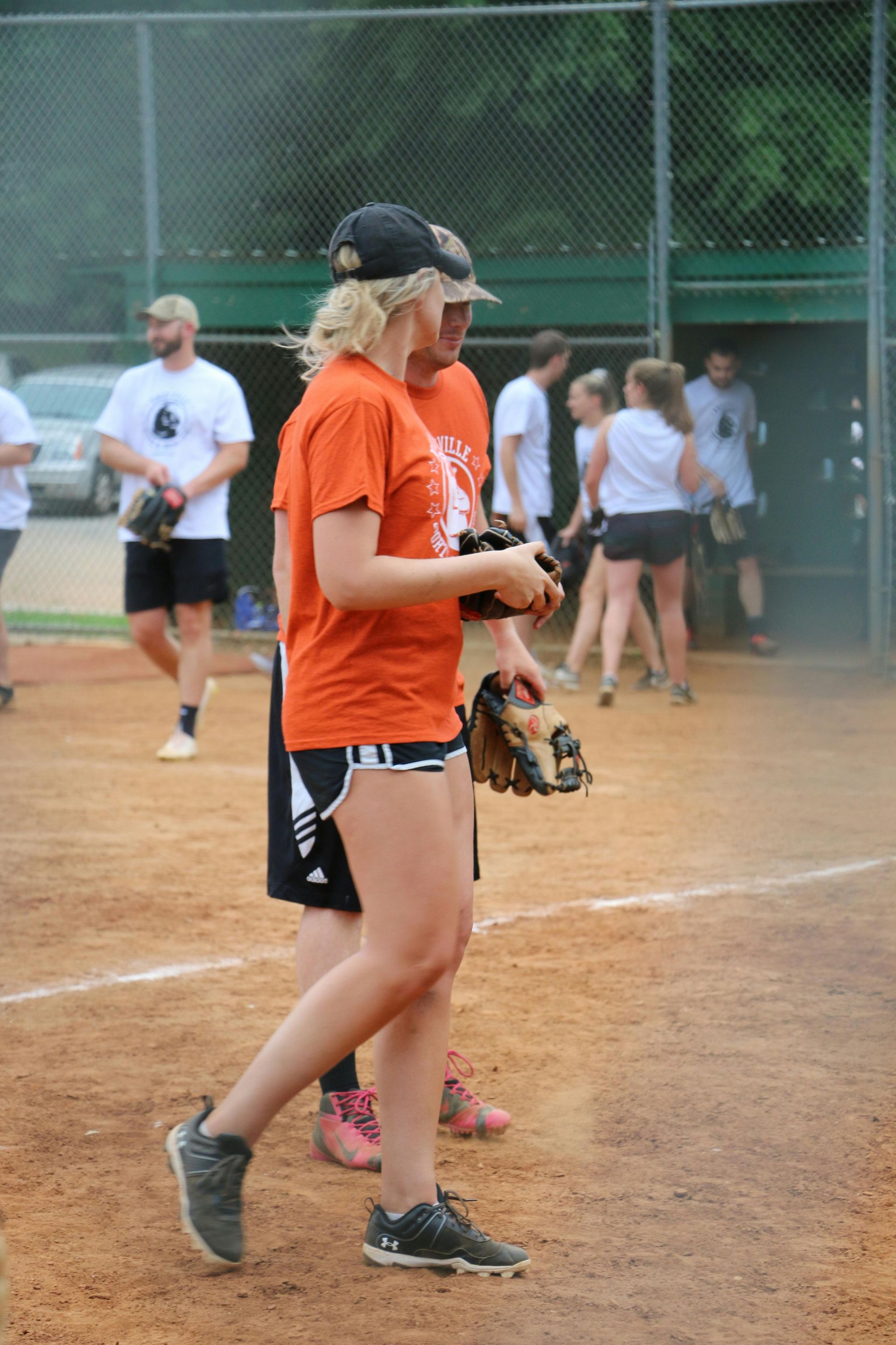 Woman in orange shirt, black shorts walks on a baseball field. Others in team shirts, background.