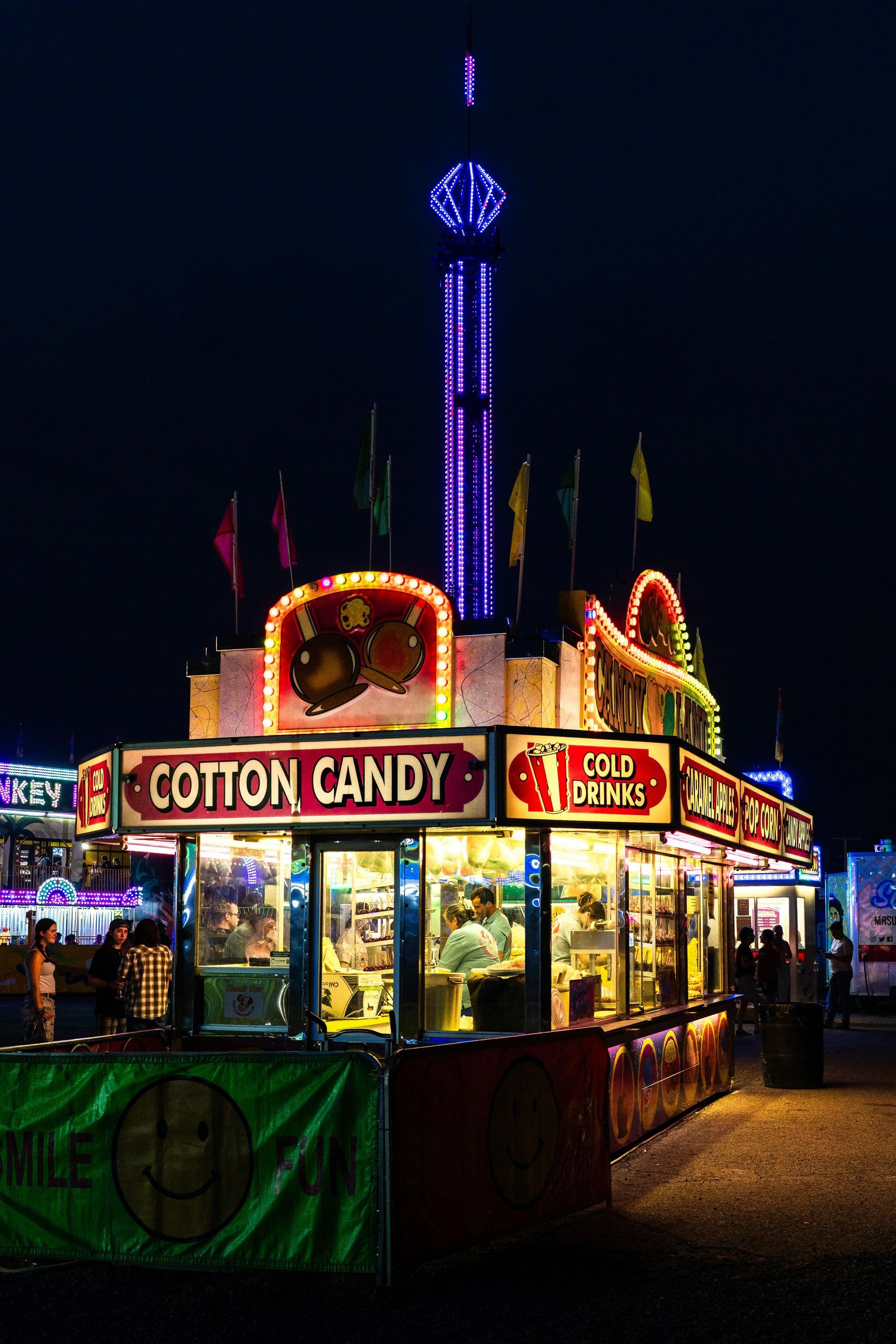 Cotton candy stand at a night carnival; illuminated sign, with tall ride in background.