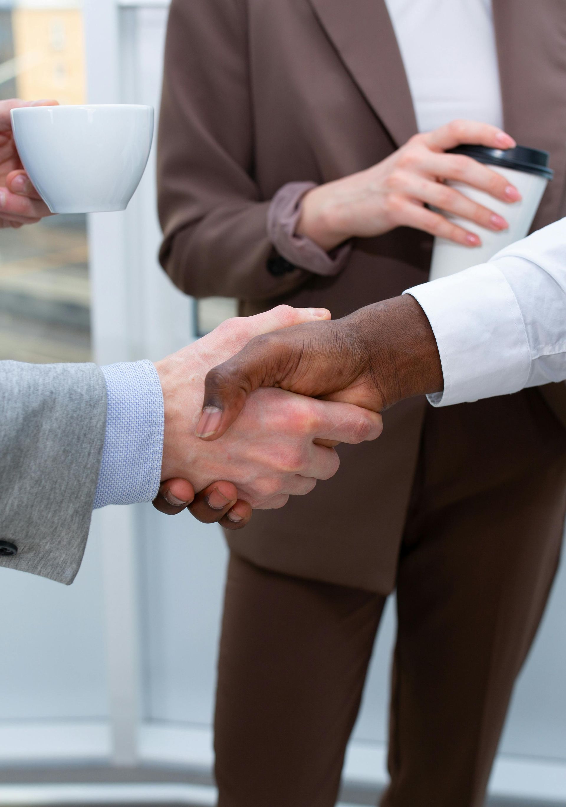 Two people in professional business attire shaking hands while holding coffee cups in an office setting.