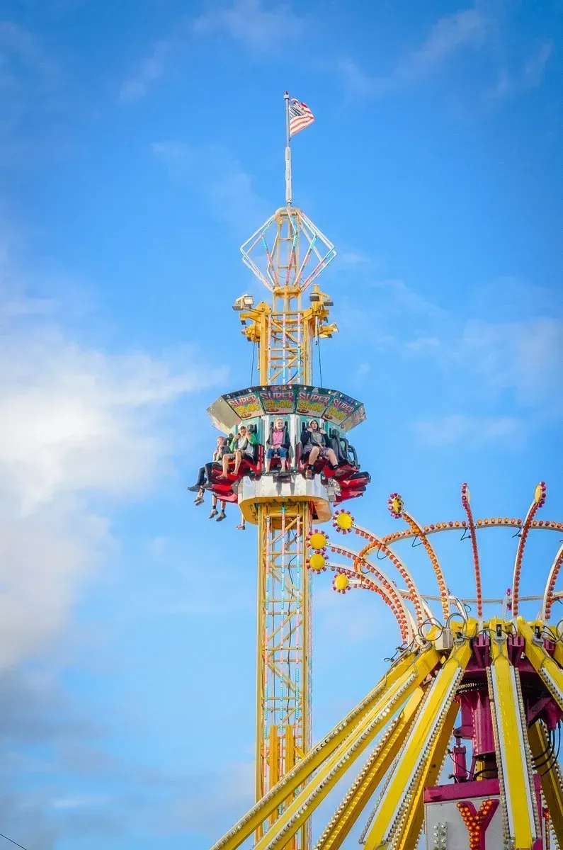 Amusement park ride with riders, against a blue sky, topped with American flag.