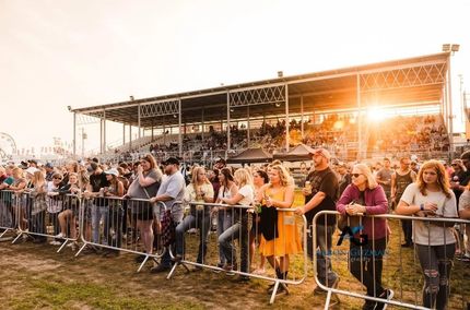 Crowd watches event behind metal barriers, with a grandstand in the background lit by sunset.