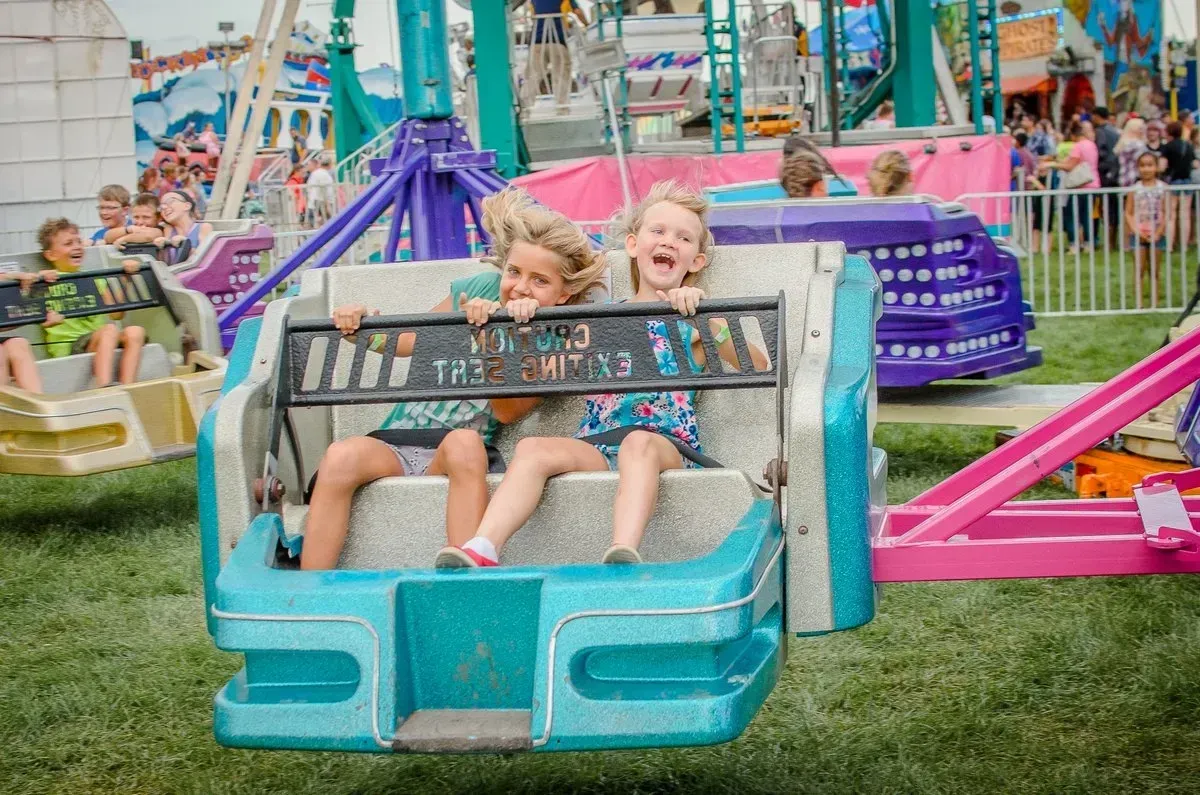 Two children laughing on a teal amusement park ride at a fair.
