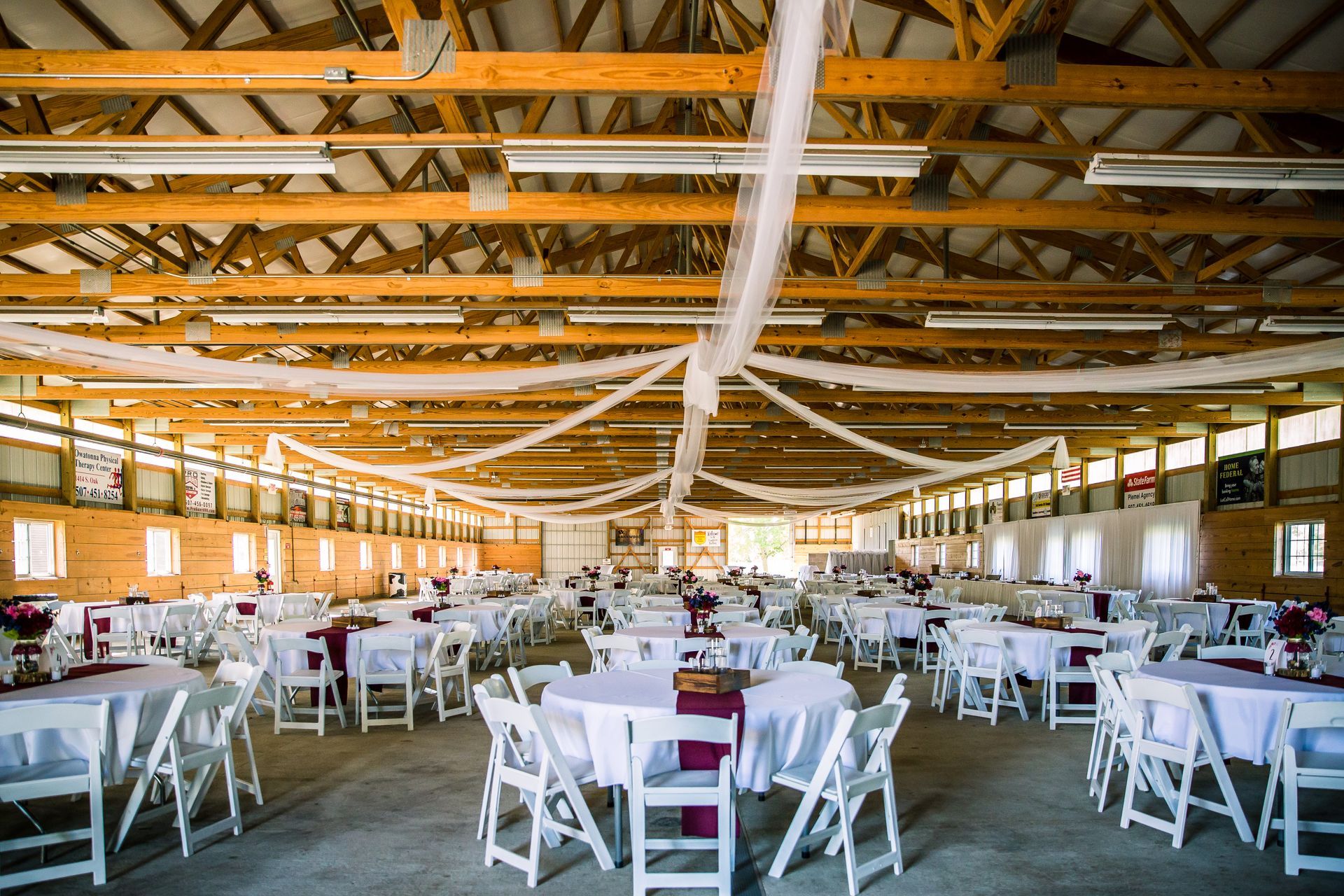 Wedding reception in a barn with round and square tables, white chairs, and decorative fabric draped from wooden beams.
